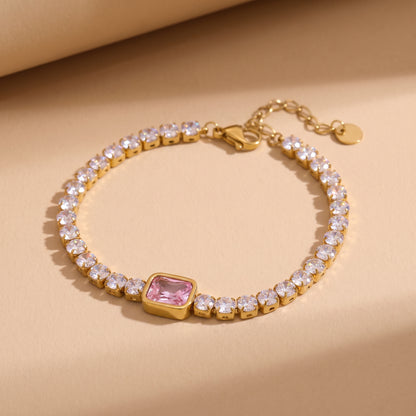 High quality Amara Gold Tennis Bracelet by Floravisha featuring clear round crystals and a center pink emerald-cut gemstone on a warm beige studio background.
