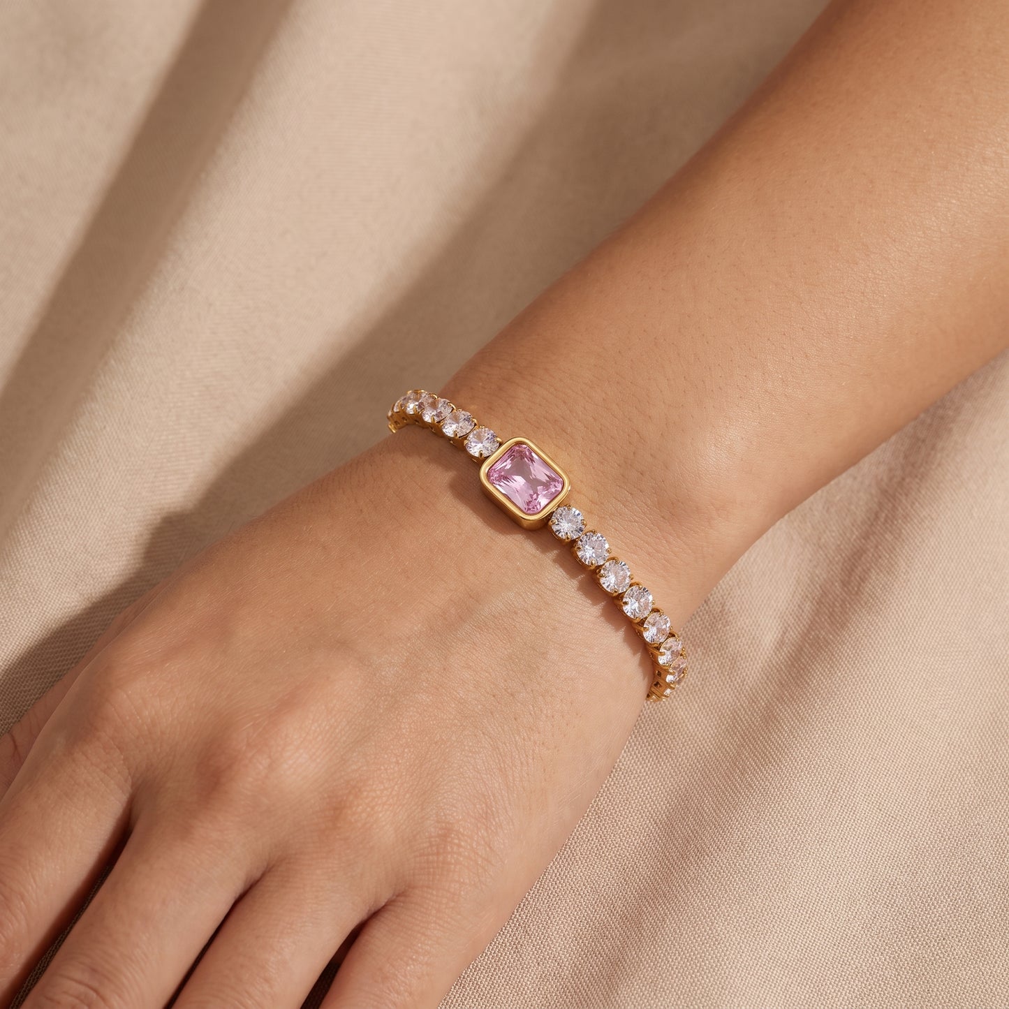 Close-up view of a woman's wrist wearing the waterproof Amara gold tennis bracelet with a central pink emerald-cut crystal against beige fabric.