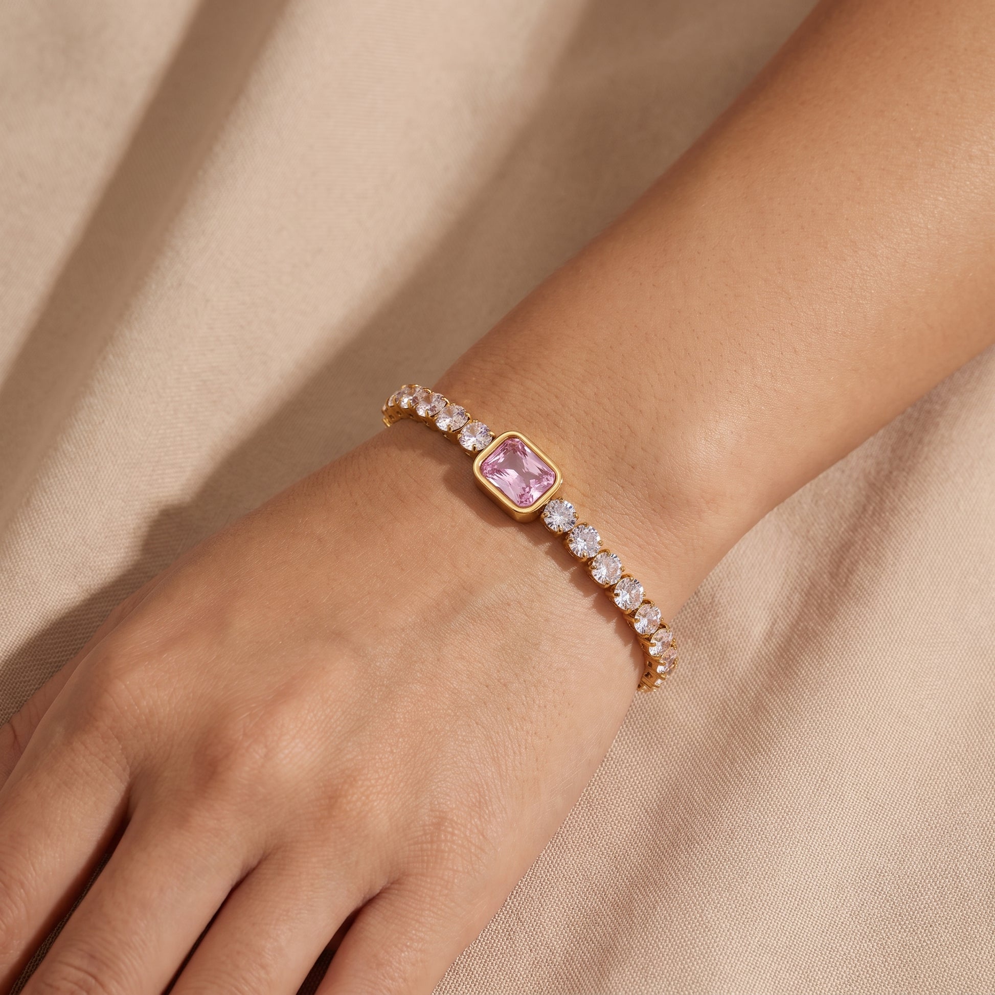 Close-up view of a woman's wrist wearing the waterproof Amara gold tennis bracelet with a central pink emerald-cut crystal against beige fabric.
