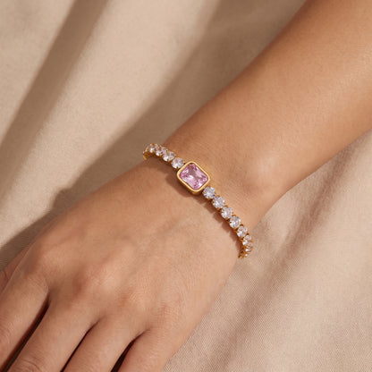 Close-up view of a woman's wrist wearing the waterproof Amara gold tennis bracelet with a central pink emerald-cut crystal against beige fabric.