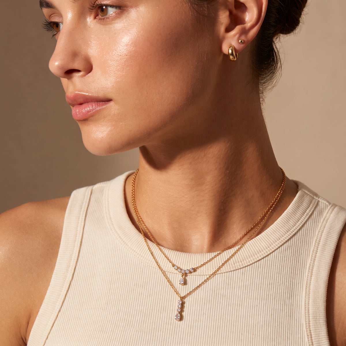 Editorial portrait of a woman wearing the waterproof Camille gold linear drop necklace layered beneath the Clarisse crystal arc necklace with a neutral ribbed top.
