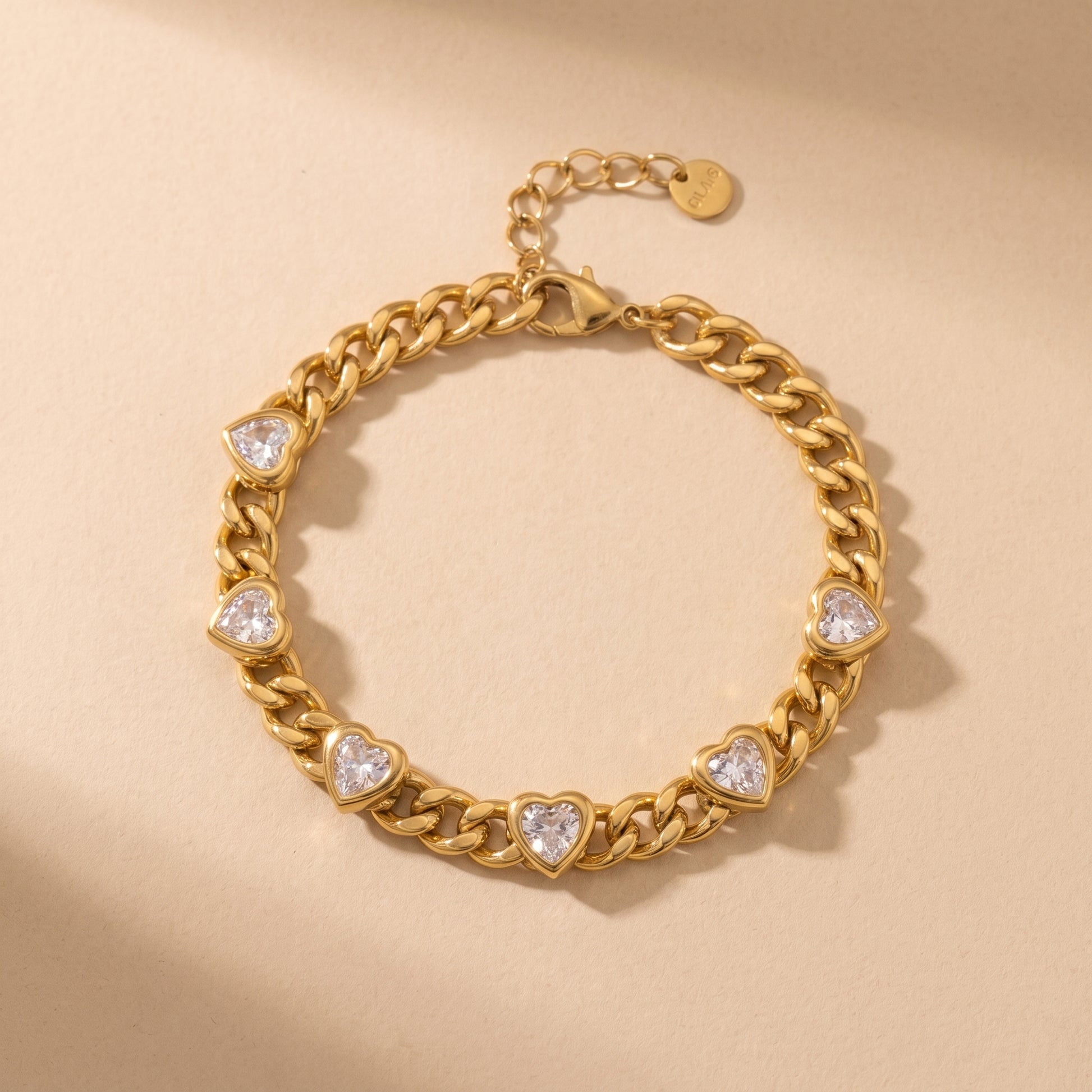 High quality Amour Gold Bracelet by Floravisha featuring five bezel-set clear heart crystals embedded along a polished curb chain resting on a warm beige studio surface.