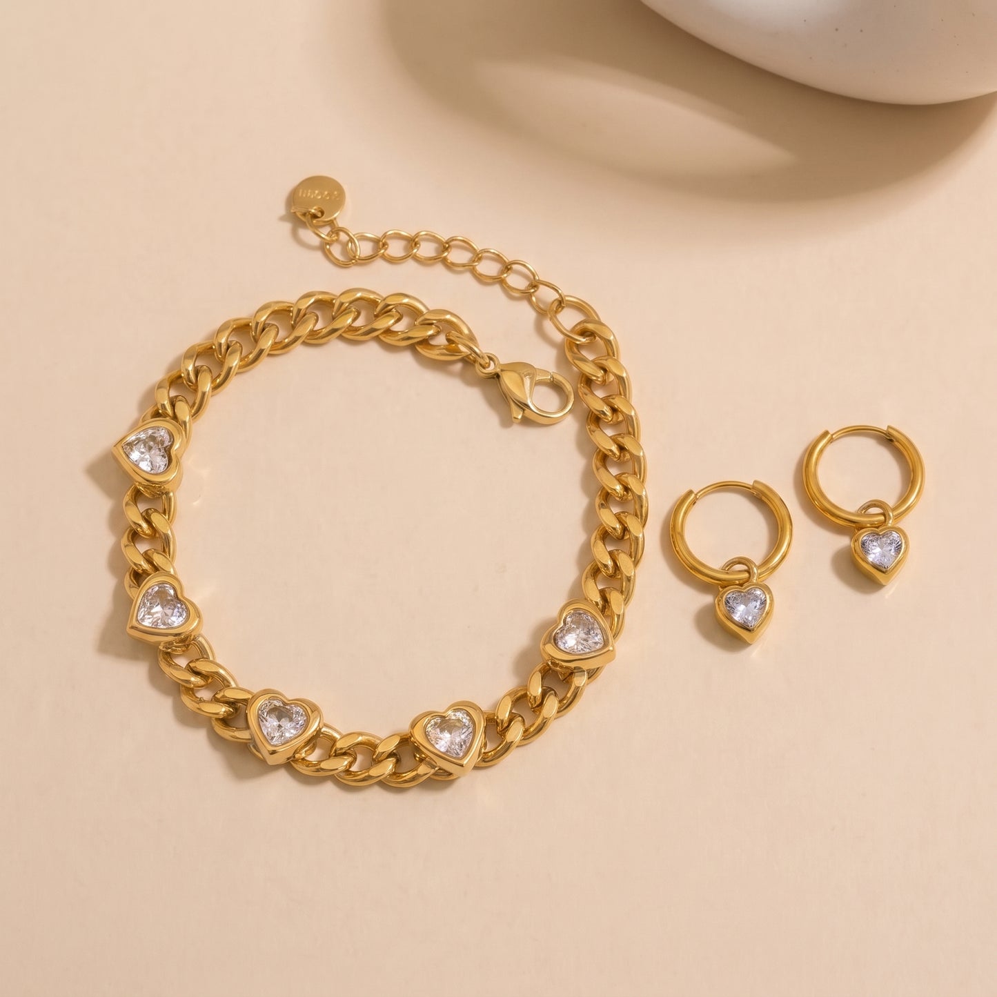 Gold chain bracelet and matching hoop earrings with heart-shaped clear stones, displayed on a warm beige background.