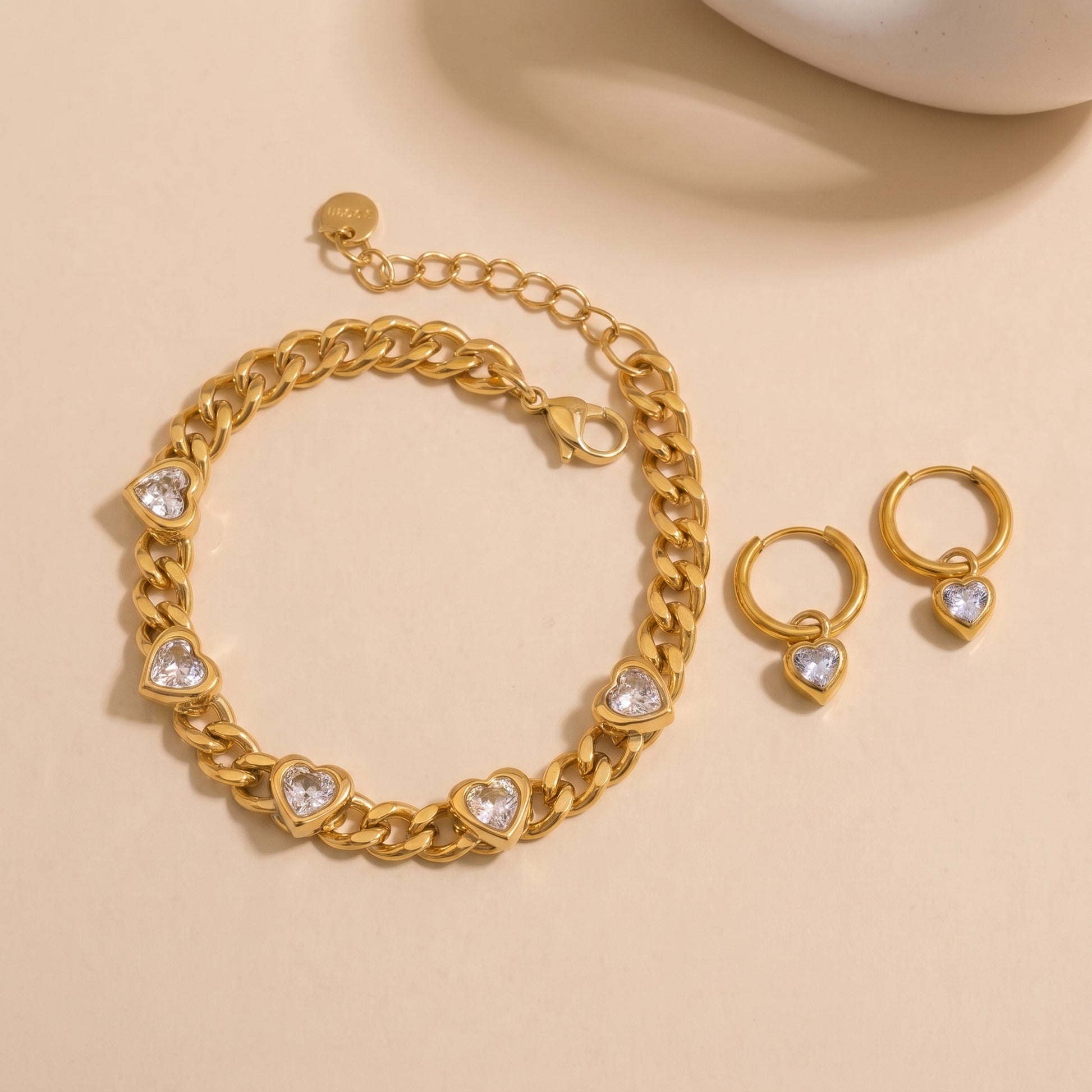 Gold chain bracelet and matching hoop earrings with heart-shaped clear stones, displayed on a warm beige background.