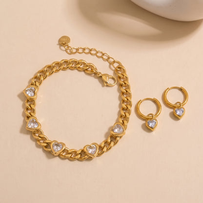 Gold chain bracelet and matching hoop earrings with heart-shaped clear stones, displayed on a warm beige background.