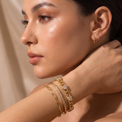 Modern gold wrist stack on a woman featuring the skin friendly Amour heart crystal curb bracelet paired with a tennis chain and sleek bangles.