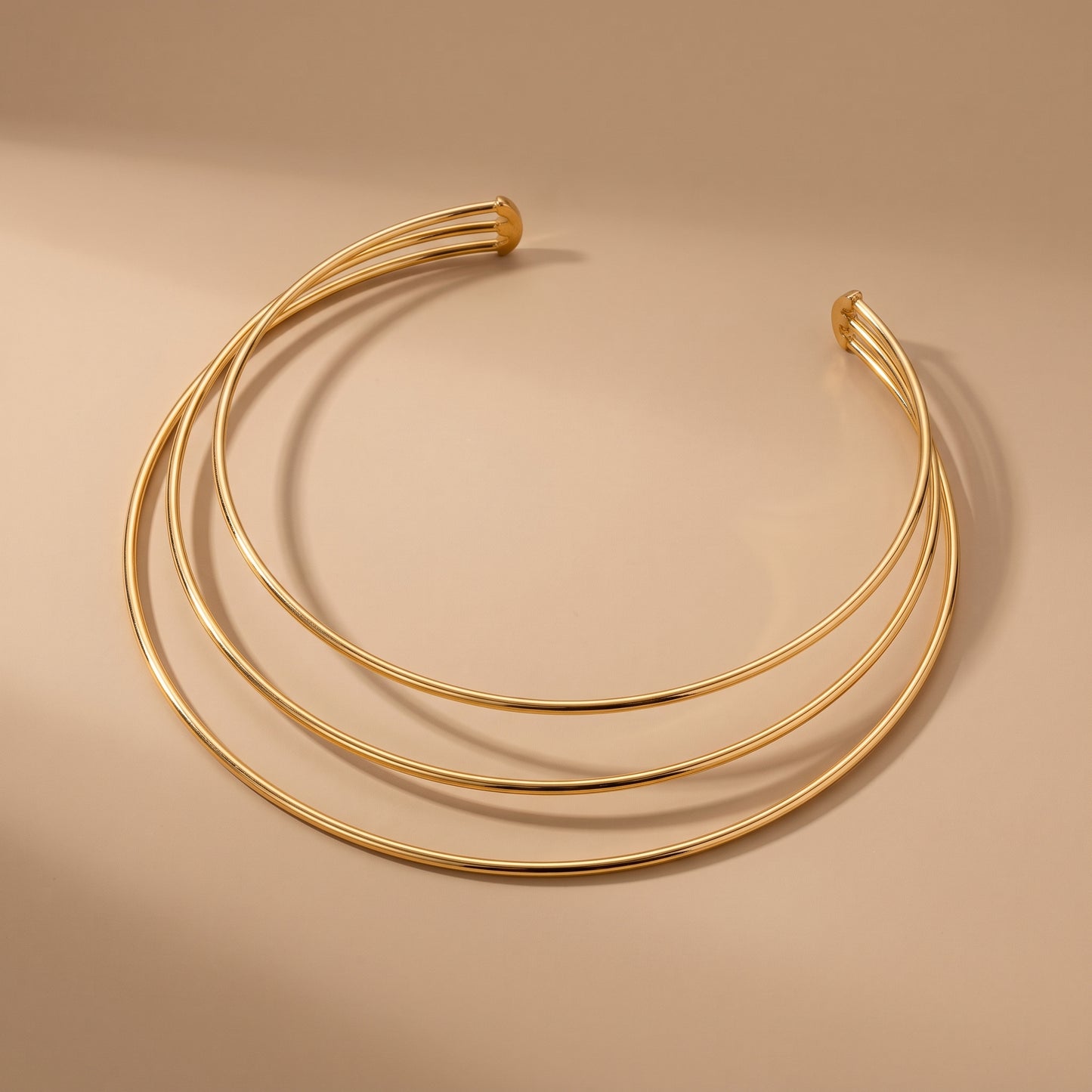 Waterproof Aura gold triple parallel band open collar choker displaying three tubular golden bands on a warm beige studio surface.