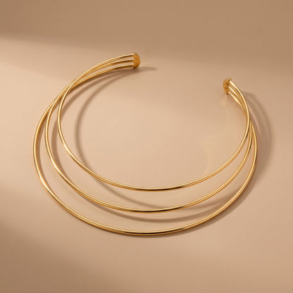 Waterproof Aura gold triple parallel band open collar choker displaying three tubular golden bands on a warm beige studio surface.
