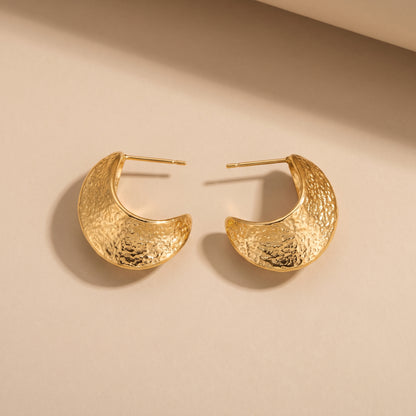 Tarnish-free Aurelia gold tapered crescent half-hoop earrings displaying hammered molten textures and stud posts on a smooth beige background.