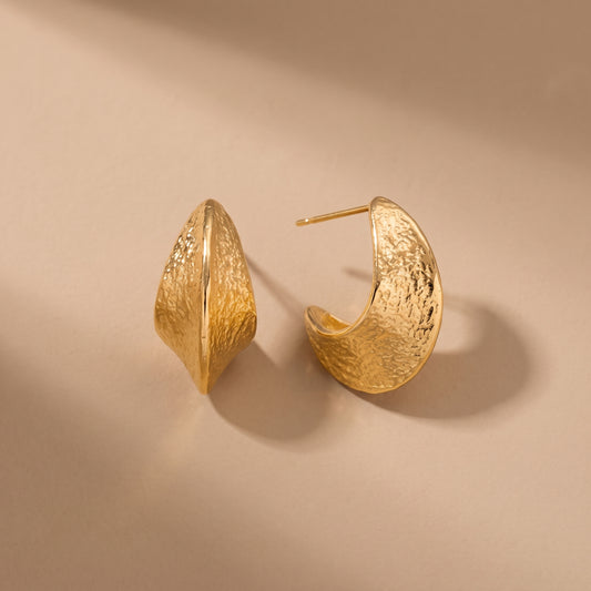 High quality Aurelia gold hammered crescent hoop earrings by Floravisha showing both the front polished spine and curved side profile on a warm beige studio surface.