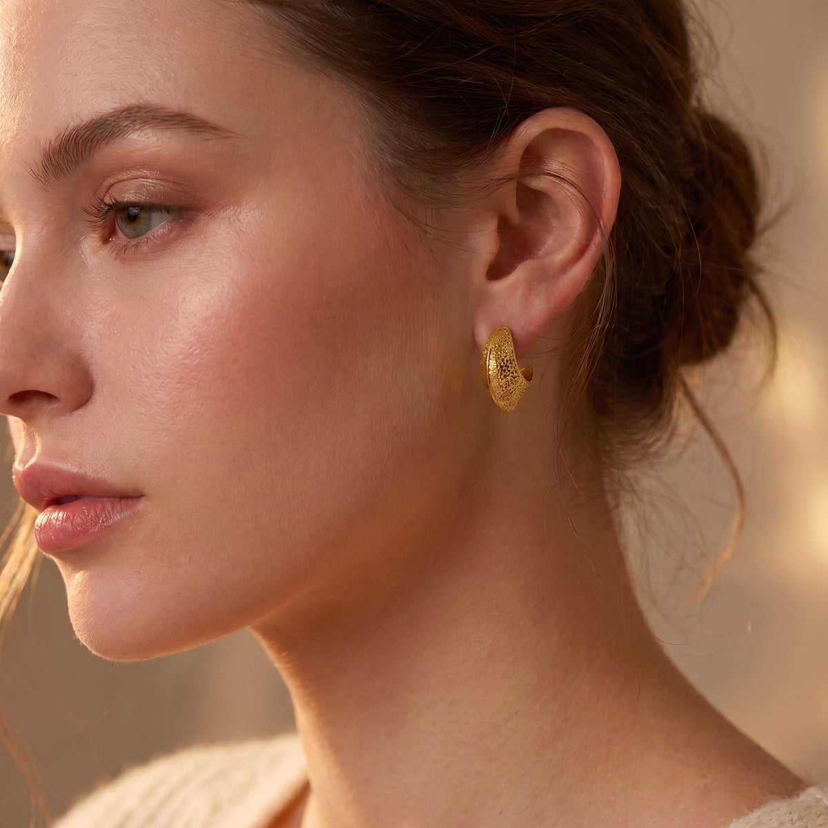 Close-up view of a woman's earlobe wearing the skin friendly Aurelia gold hammered crescent hoop earring paired with a cream knit sweater in natural warm light.