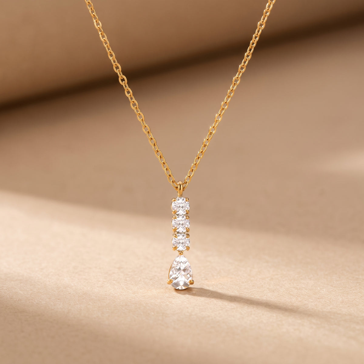 High quality Camille gold linear necklace displaying a vertical column of three round crystals and a suspended pear-cut teardrop pendant on a warm beige studio surface.