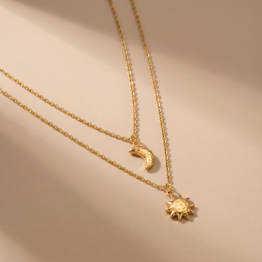 Tarnish-free Céleste gold layered necklace featuring a hammered crescent moon and sunburst pendant resting on a warm beige background in natural light.