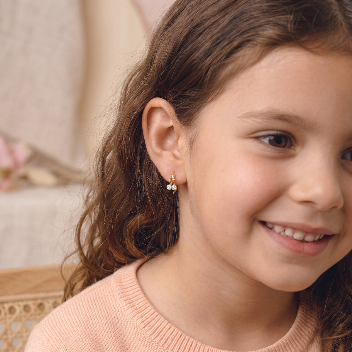 Smiling young girl wearing the lightweight, hypoallergenic Cerise gold pavé cherry huggie hoop earrings with a peach knit sweater.