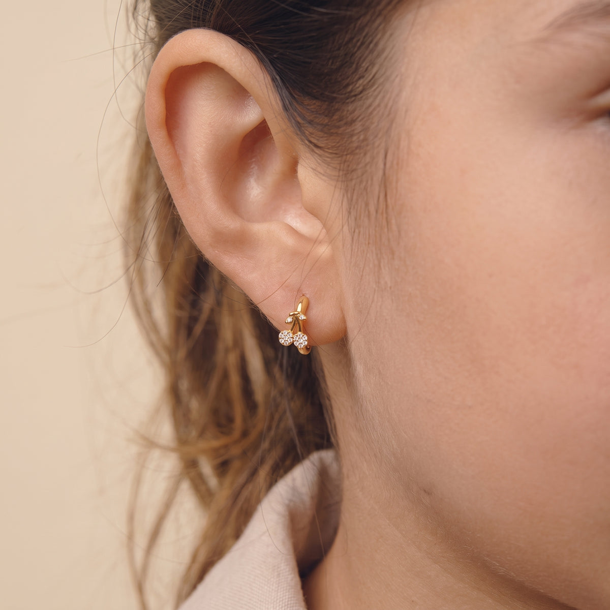Close-up view of a woman's earlobe wearing the skin friendly Cerise gold pavé cherry huggie hoop earring in soft natural lighting.