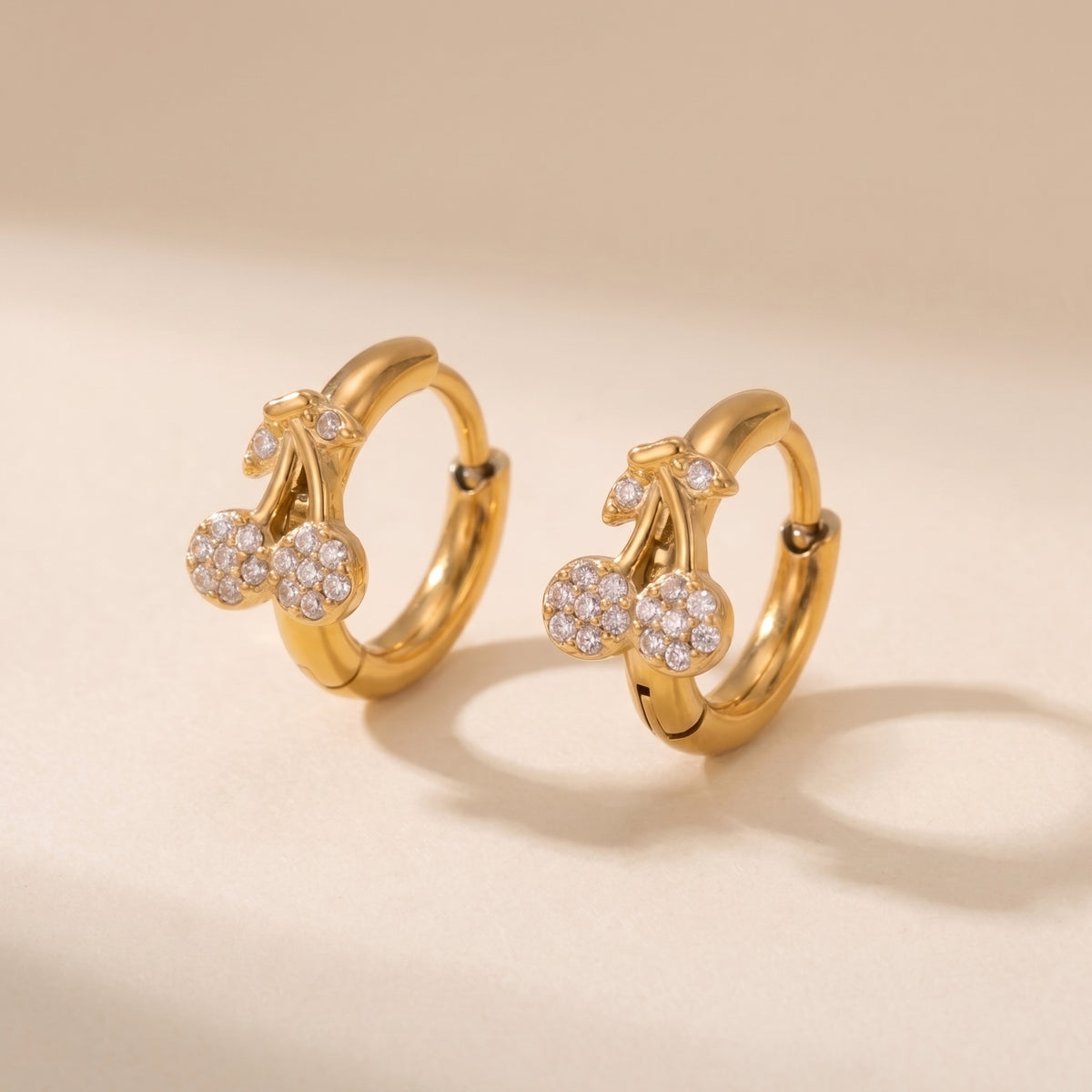 High quality Cerise gold pavé cherry huggie hoop earrings by Floravisha showing clear cubic zirconia crystals and click-hinge clasps on a warm beige background.