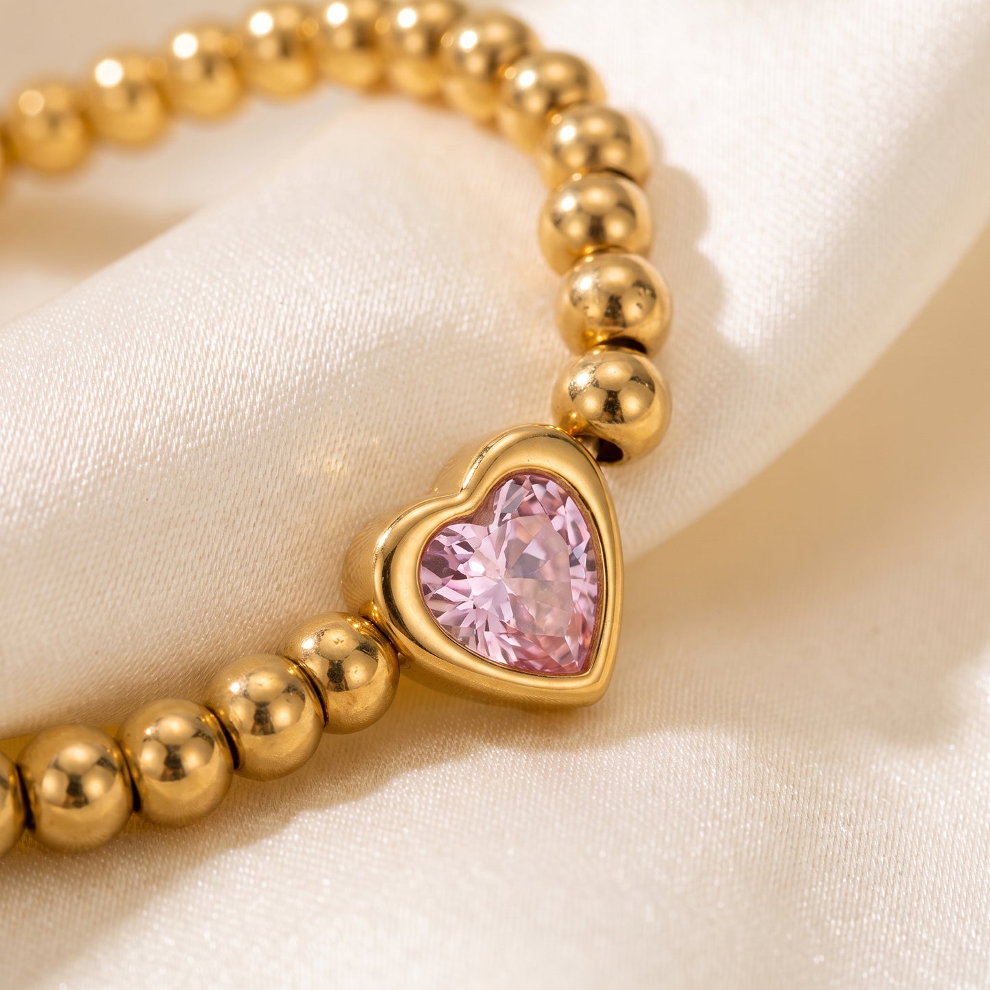 Extreme macro close-up view of the tarnish-free Chérie gold bead bracelet showcasing the bezel-set pink heart crystal on draped champagne silk fabric.