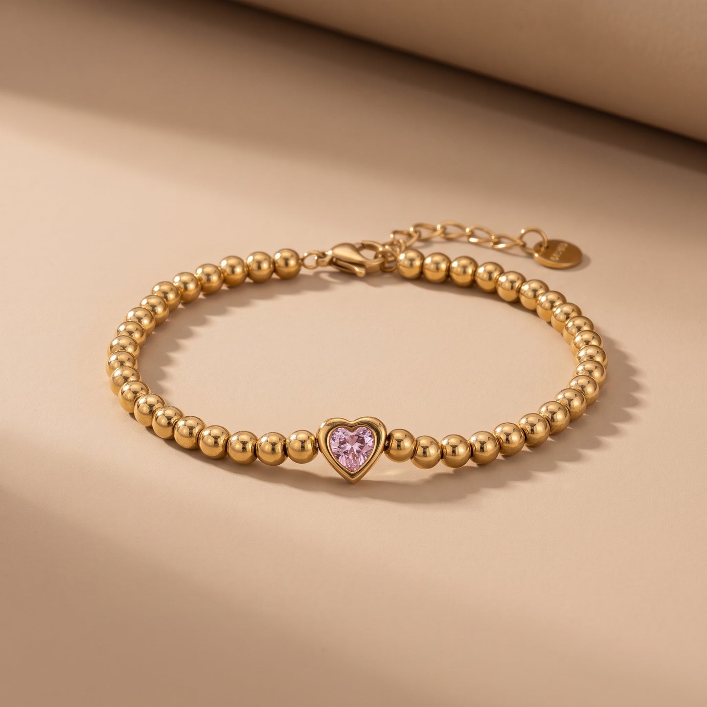 High quality Chérie Gold Bracelet by Floravisha featuring polished golden ball beads and a central pink heart crystal on a warm beige studio background.