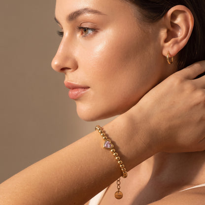Close-up view of a woman's wrist wearing the tarnish-free Chérie gold bead bracelet with central pink heart gem in natural lighting.
