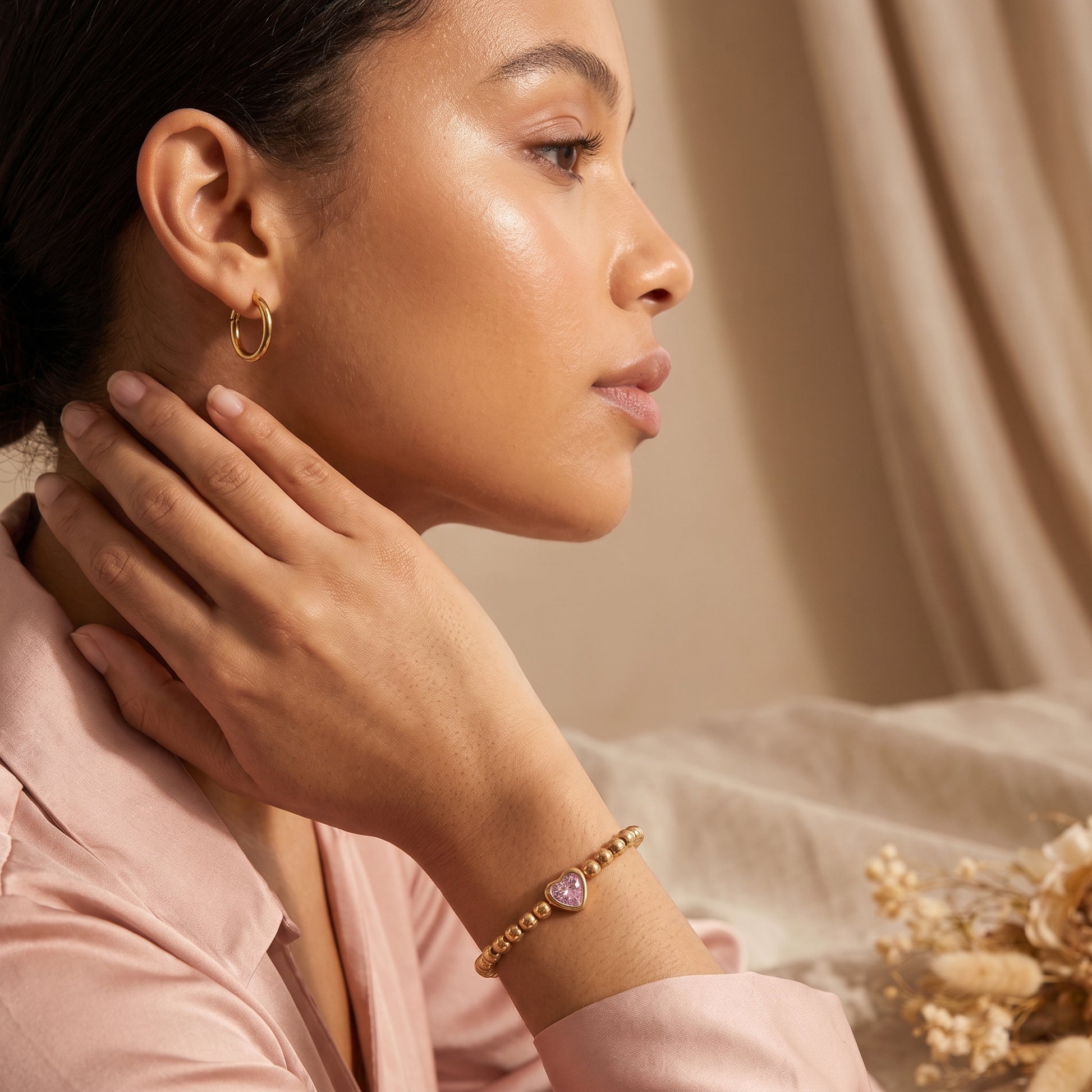 Side profile of a woman wearing the skin friendly Chérie gold pink heart bead bracelet paired with a pink silk shirt in warm studio lighting.