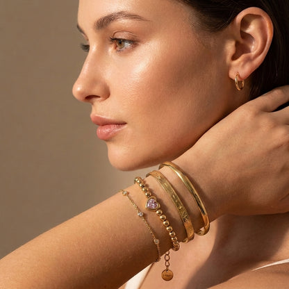 Modern gold wrist stack on a woman featuring the waterproof Chérie pink heart bead bracelet layered with the Astrid engraved cuff and Aura sleek cuff.