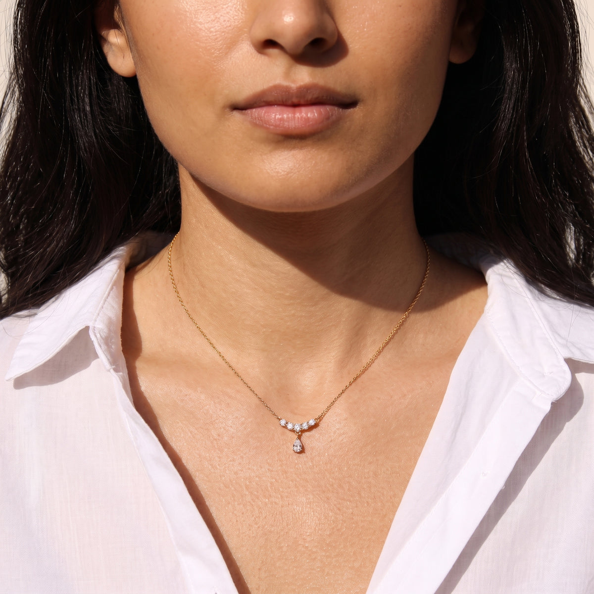 Close-up view of a woman's collarbone wearing the skin friendly Clarisse gold crystal arc teardrop necklace with an open white linen shirt in natural sunlight.