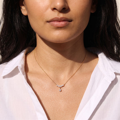 Close-up view of a woman's collarbone wearing the skin friendly Clarisse gold crystal arc teardrop necklace with an open white linen shirt in natural sunlight.
