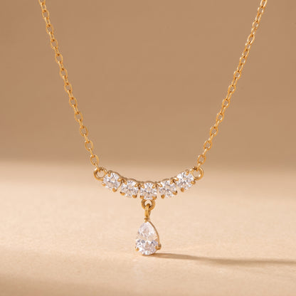 High quality Clarisse gold necklace displaying a contoured 5-stone round crystal arc and suspended pear-cut teardrop pendant on a warm beige background.