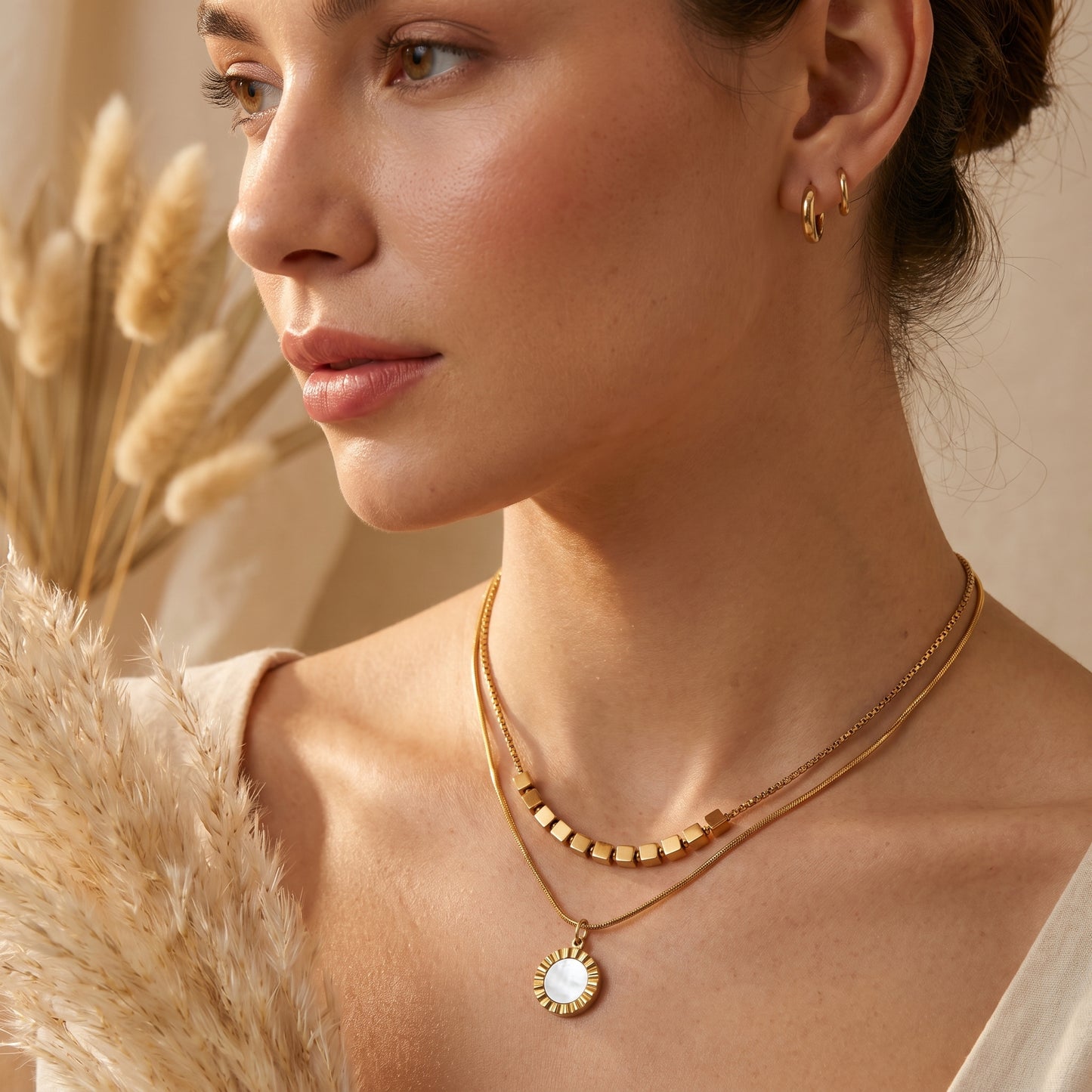 Woman wearing gold necklaces and earrings. High quality waterproof gold plated double layer necklace featuring gold cube accents and a black natural stone pendant on a beige background