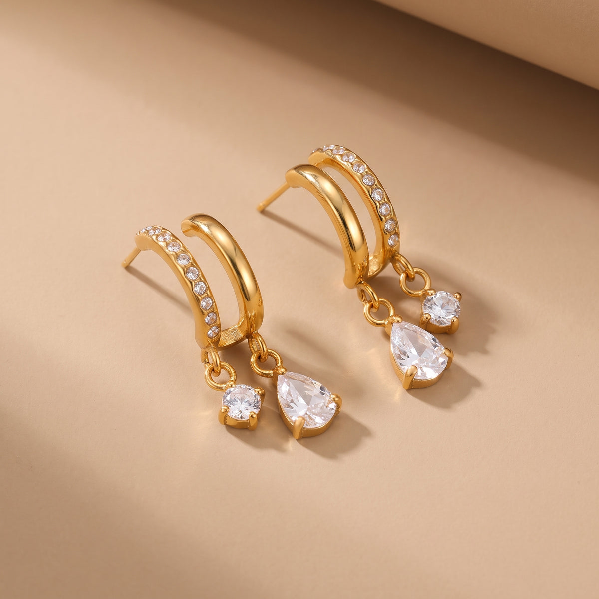 High quality Delphine Gold Drop Earrings by Floravisha featuring a dual arch of smooth gold and pavé crystals with dangling teardrop gems on a warm beige studio surface.