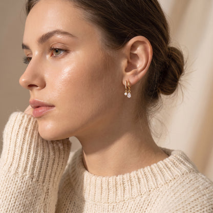 Side profile of a woman wearing the waterproof Delphine gold teardrop crystal drop earrings paired with an off-white knit sweater in soft studio lighting.