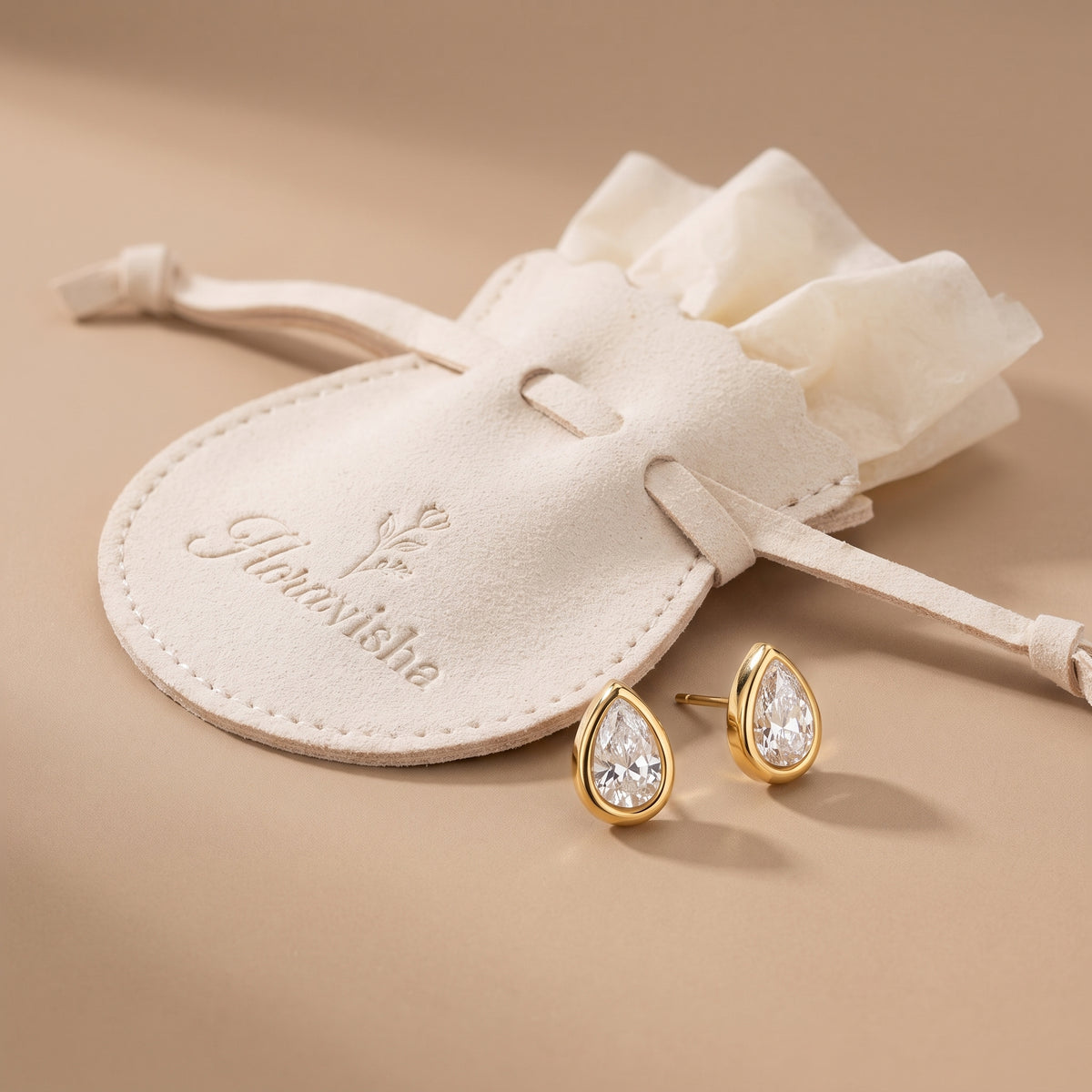 Pair of waterproof Delphine Petite gold teardrop stud earrings resting next to an off-white Floravisha scalloped suede jewelry pouch.