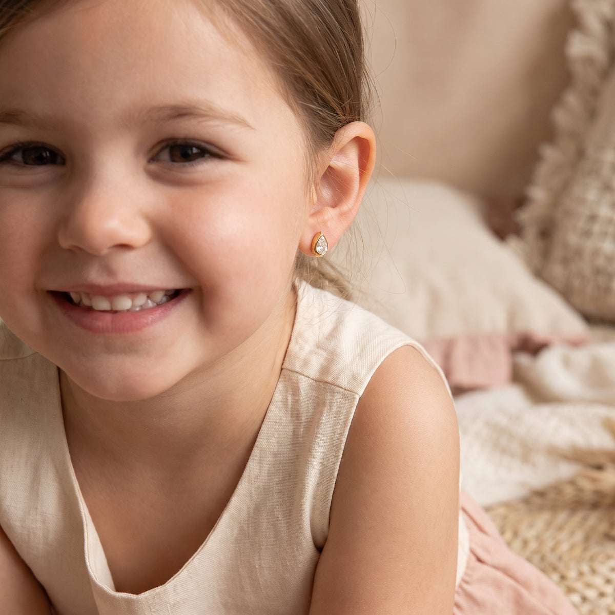 Close-up of a smiling girl with brown wavy hair wearing the lightweight, skin friendly Delphine Petite gold teardrop solitaire stud earrings.