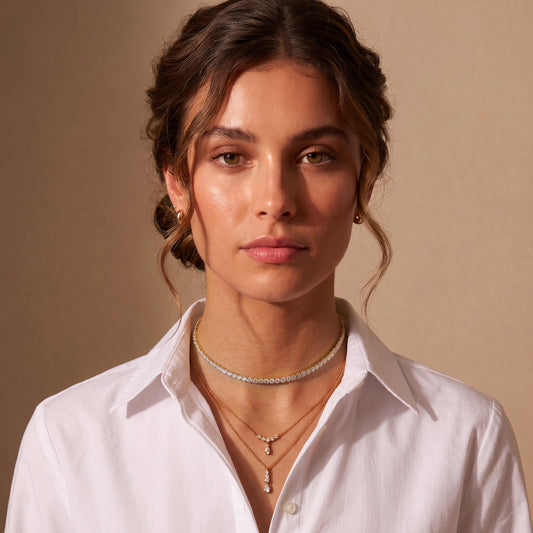 Close-up view of a woman wearing the waterproof Étoile gold tennis choker layered with the Clarisse crystal arc and Camille drop necklaces with a white button-down shirt.