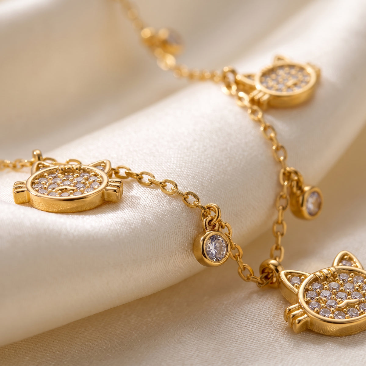 Extreme macro close-up of the tarnish-free Féline gold necklace showing micro-pavé crystal cat charms and bezel-set round gems on draped ivory silk.