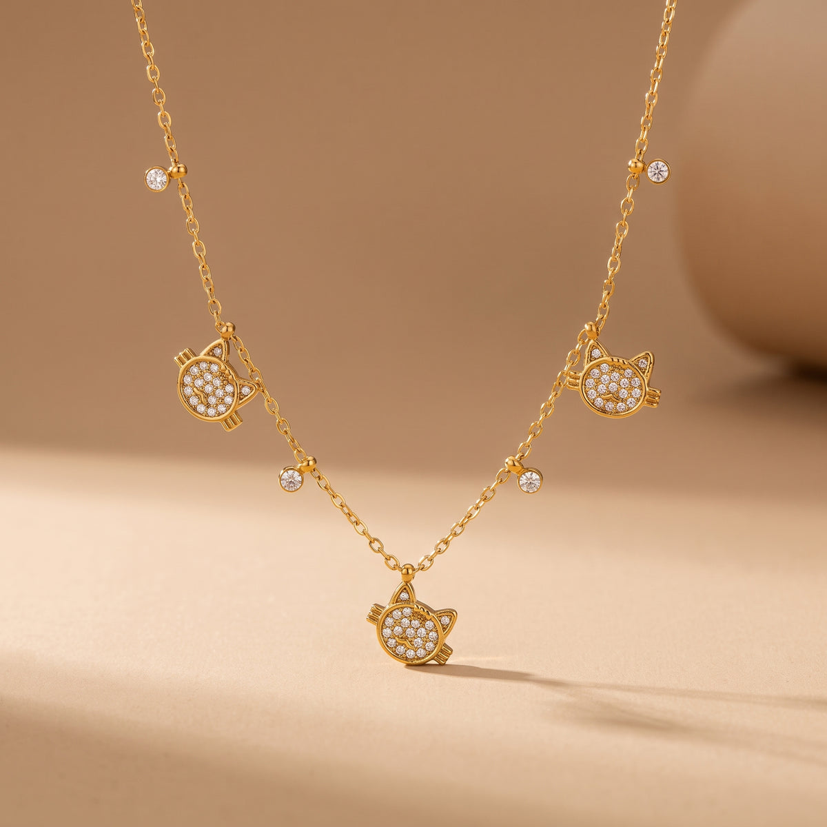 High quality Féline gold pavé cat necklace by Floravisha displaying three crystal cat charms and dangling bezel drops suspended on a warm beige background.