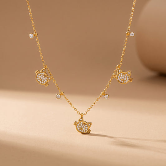 High quality Féline gold pavé cat necklace by Floravisha displaying three crystal cat charms and dangling bezel drops suspended on a warm beige background.