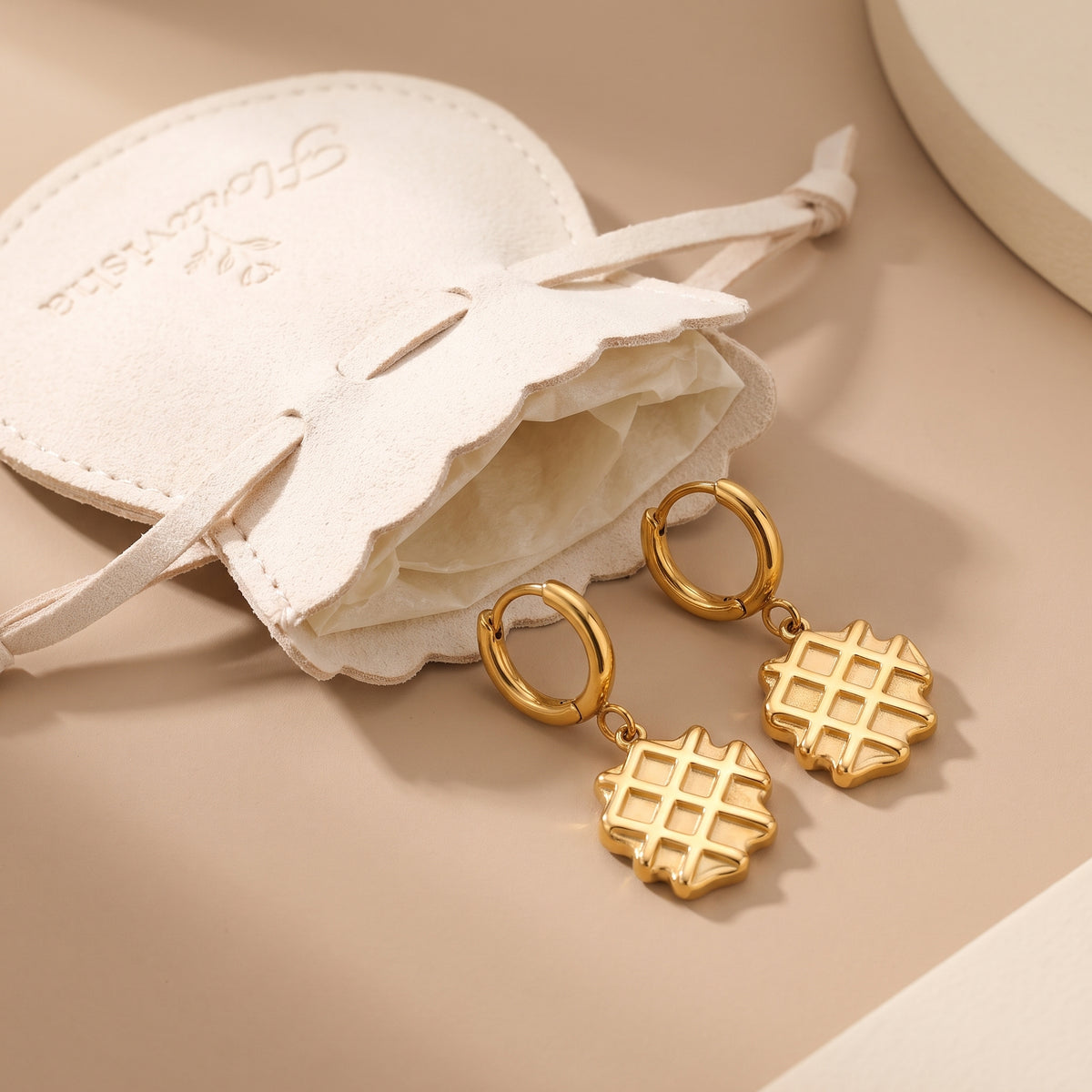 High quality La Gaufre gold waffle charm huggie hoop earrings resting next to an off-white Floravisha scalloped suede jewelry pouch.