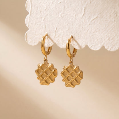 Pair of waterproof La Gaufre gold waffle charm huggie earrings suspended from a scalloped white display card showing grid texture.