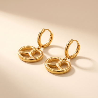 High quality Le Bretzel gold pretzel charm huggie hoop earrings by Floravisha showing openwork knotted charms and click-hinge clasps on a warm beige surface.