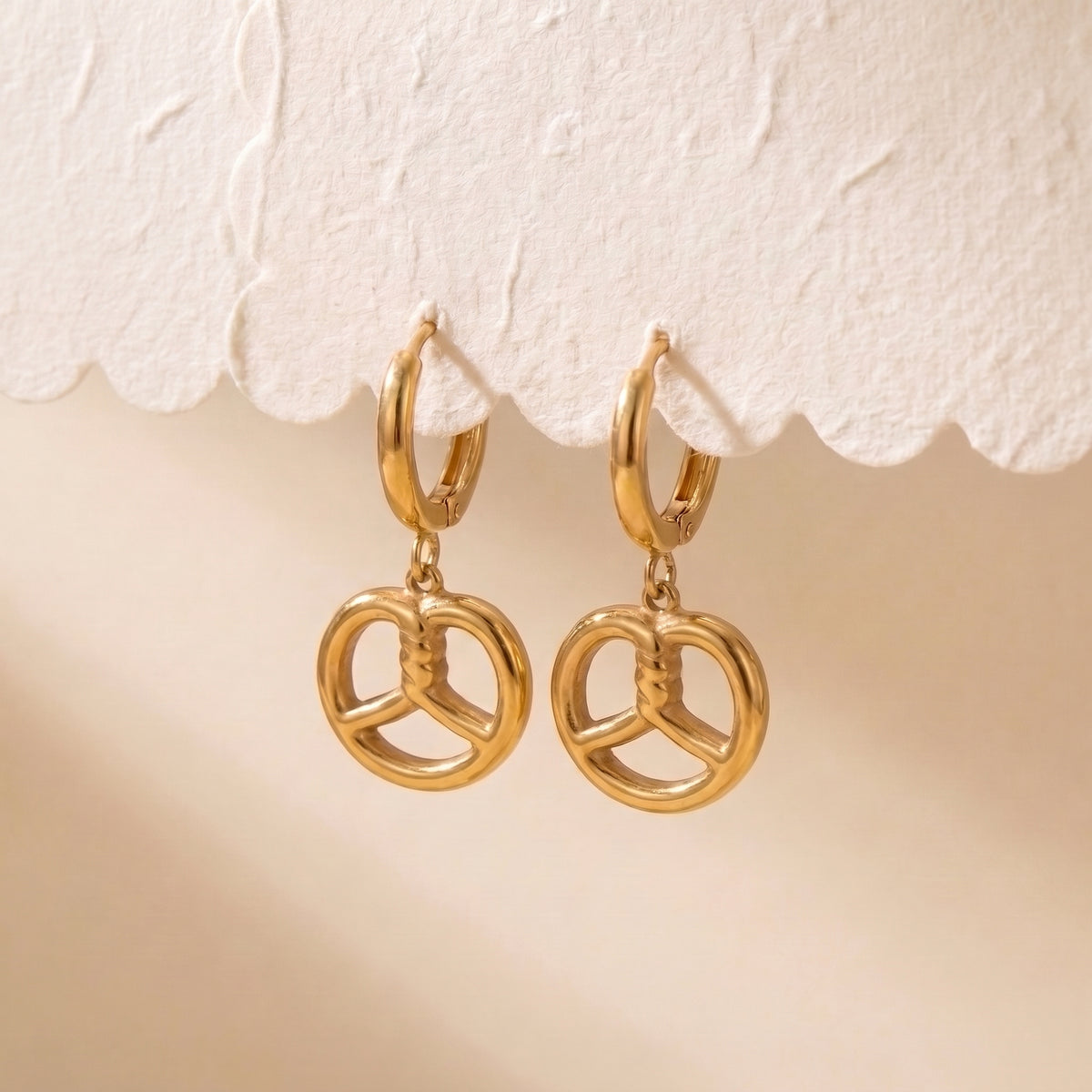 Tarnish-free Le Bretzel gold pretzel charm earrings suspended from a textured scalloped display card showing balanced drop alignment.