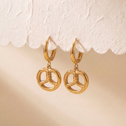 Tarnish-free Le Bretzel gold pretzel charm earrings suspended from a textured scalloped display card showing balanced drop alignment.