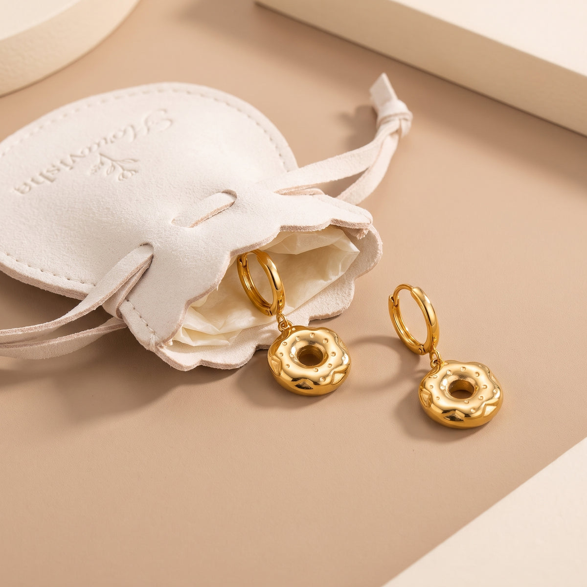 Tarnish-free Le Donut gold frosted donut charm huggie earrings resting beside an off-white Floravisha scalloped suede drawstring jewelry gift pouch.