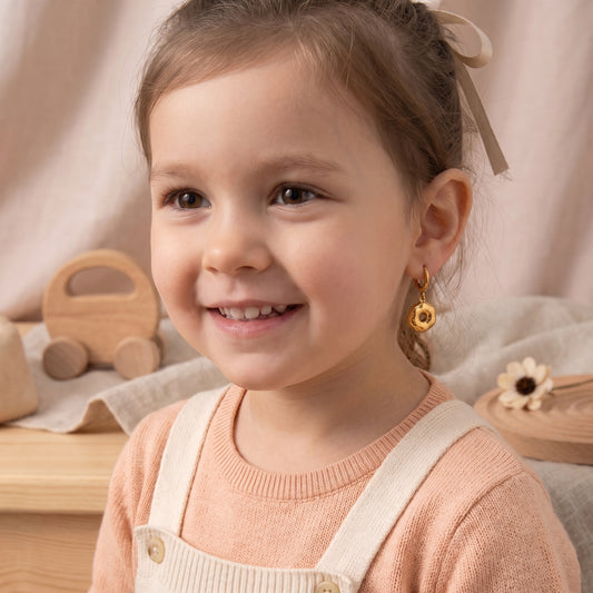 Close-up of a smiling young girl wearing the lightweight, hypoallergenic Le Donut gold frosted donut charm huggie earrings with a peach knit top.
