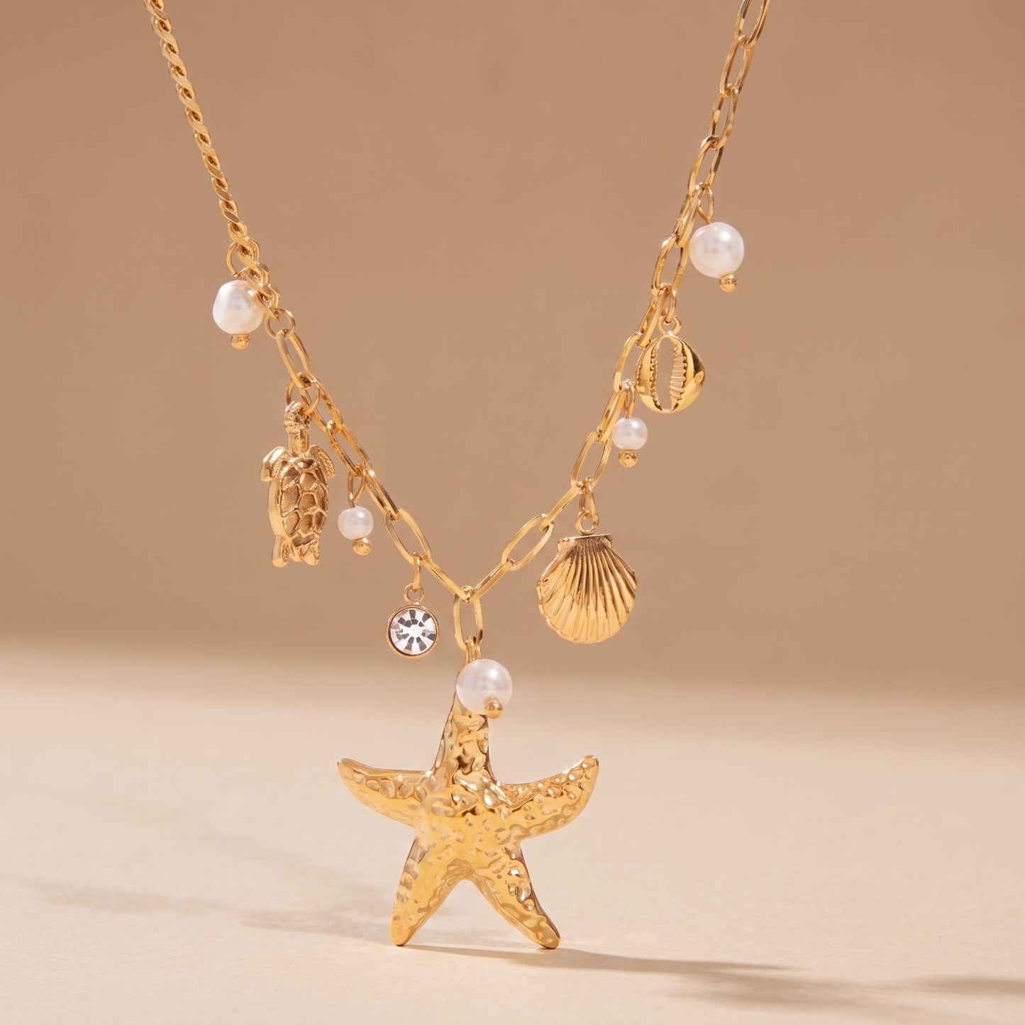High quality Oceane Gold Necklace by Floravisha featuring a gold starfish, sea turtle, and pearl charms on a warm beige background in direct sunlight.oceane-gold-coastal-starfish-necklace-studio-flatlay.jpg