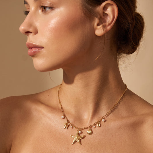 Skin friendly Oceane gold coastal charm necklace with starfish, sea turtle, and pearl drops on a model's collarbone in warm natural lighting.