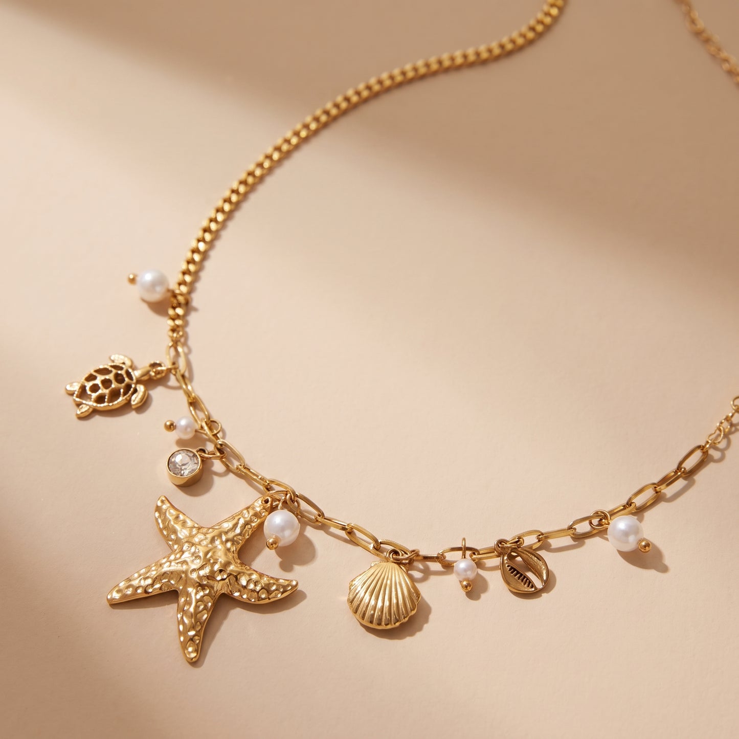 Tarnish-free Oceane gold coastal charm necklace with starfish, scallop shell, and pearl accents resting on a smooth beige travertine surface.