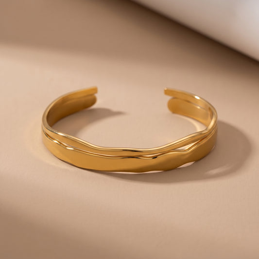 Waterproof Double Layered Ondine gold molten wave open cuff bangle resting on a warm beige studio surface.