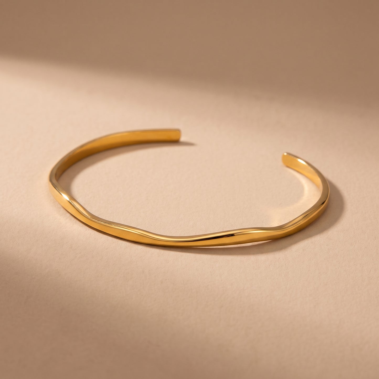 High quality delicate Slim Ondine gold undulating wave open cuff bangle resting on a warm beige studio surface.