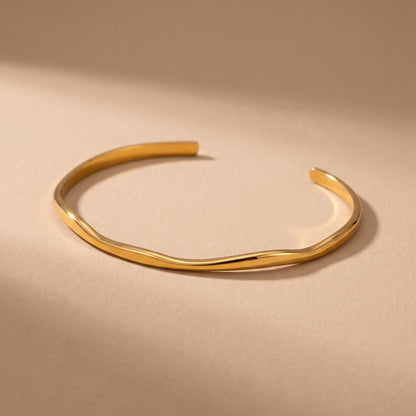 High quality delicate Slim Ondine gold undulating wave open cuff bangle resting on a warm beige studio surface.